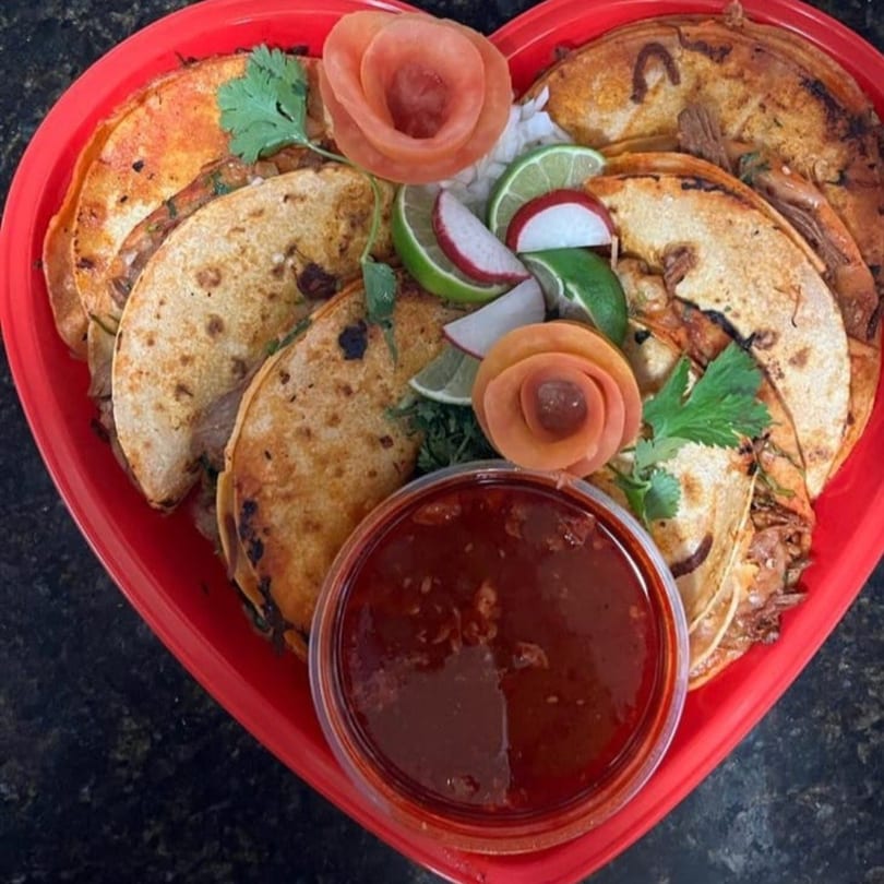Heart-shaped platter of Taqueria Los Arcos birria tacos with lime, radish, cilantro, and dipping sauce.