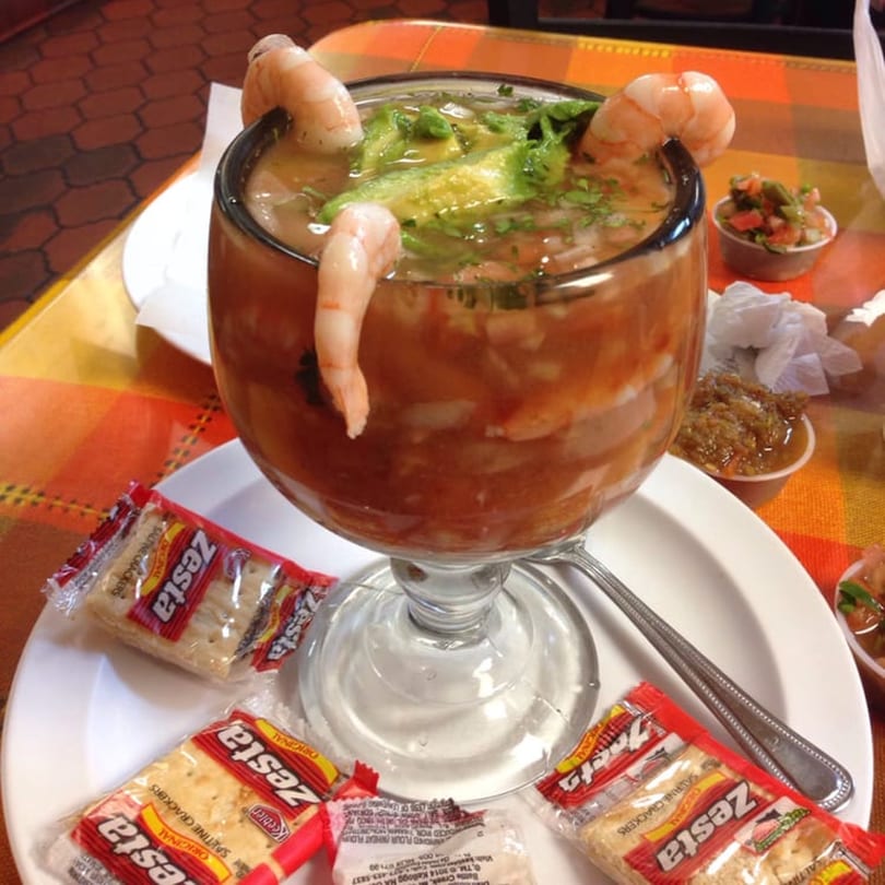 Shrimp cocktail with avocado and cilantro in a large glass, served with crackers and salsa.