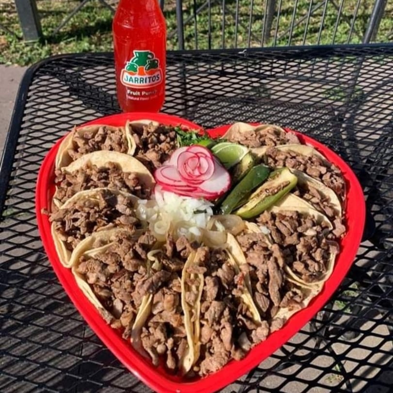 A heart-shaped platter of carne asada tacos with radishes, onions, jalapeños, and a Jarritos soda.