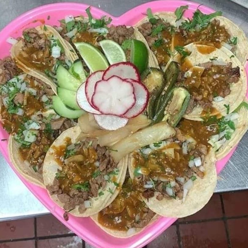 A heart-shaped pink platter filled with many tacos, garnished with radishes, limes, and cucumbers.