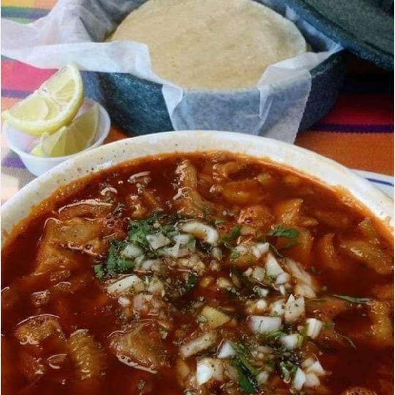 A bowl of menudo soup with chopped onions and cilantro, alongside a basket of tortillas and lemon wedges.